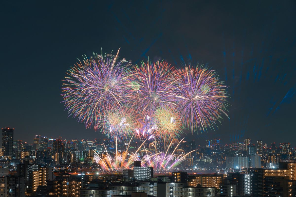 Colorful fireworks exploding above a river as people in yukata watch from the riverbank during a Japanese summer festival.