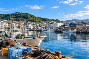 Traditional Japanese harbor town with fishing boats, waterfront buildings, and calm blue water on a sunny morning